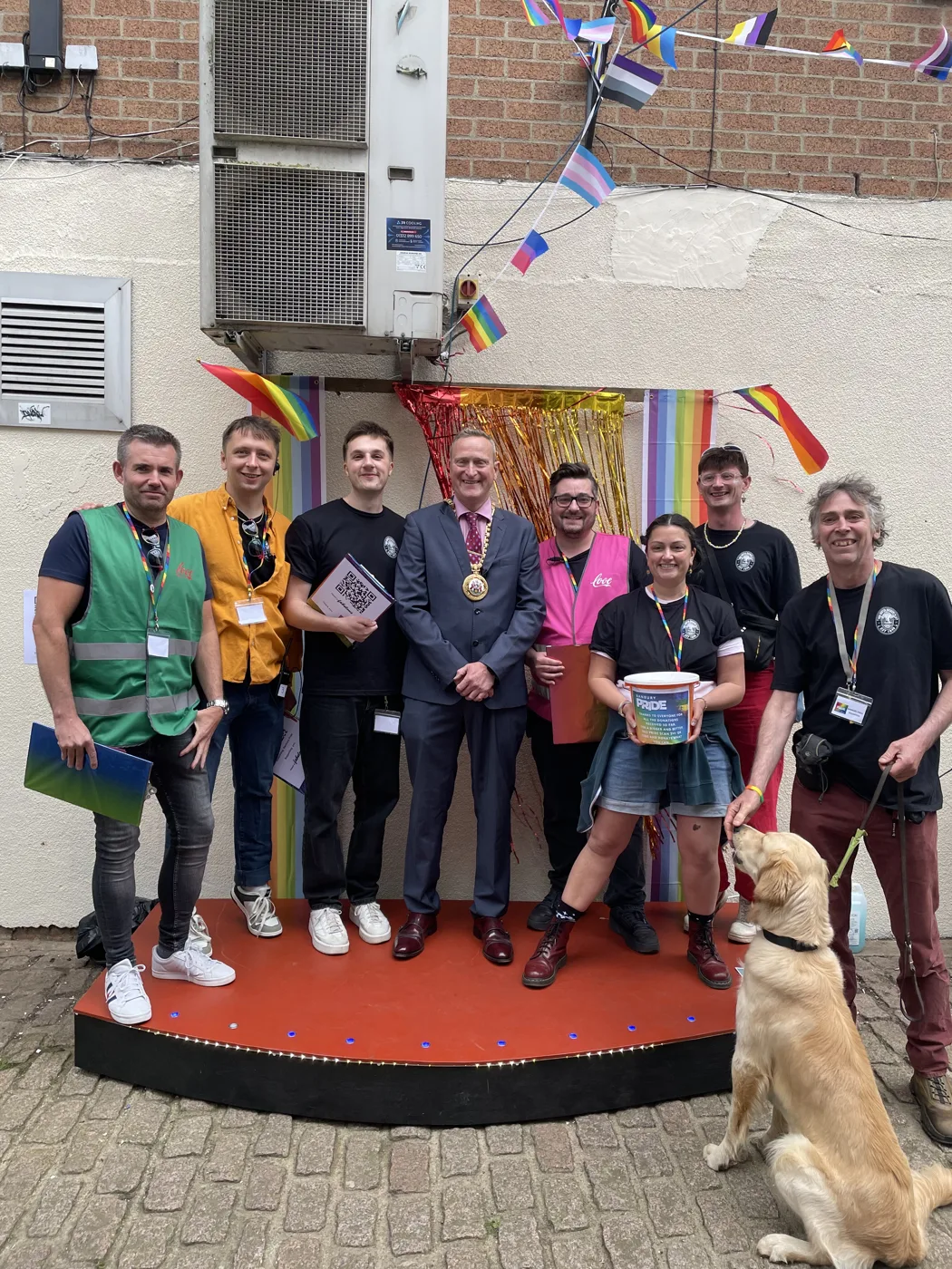 Eight people and a golden retriever on a small stage decorated with rainbow and Pride flags, one man in the centre wearing a mayoral chain