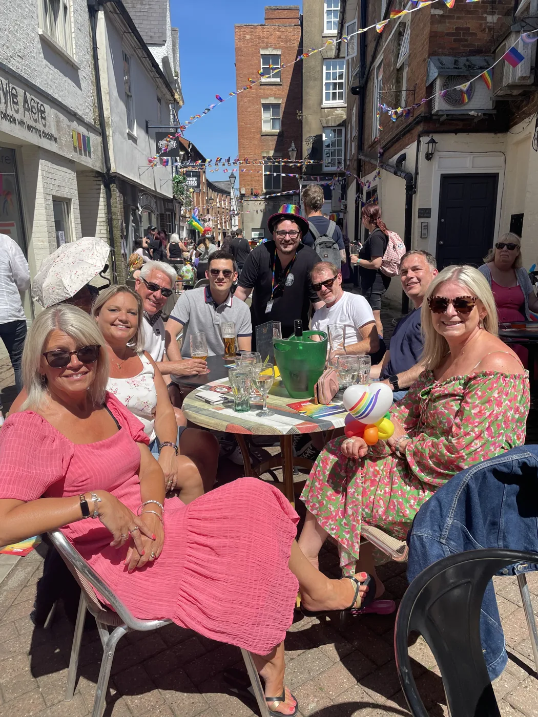 Friends sitting round an outdoor table on a sunny street hung with rainbow bunting, with drinks and a rainbow balloon