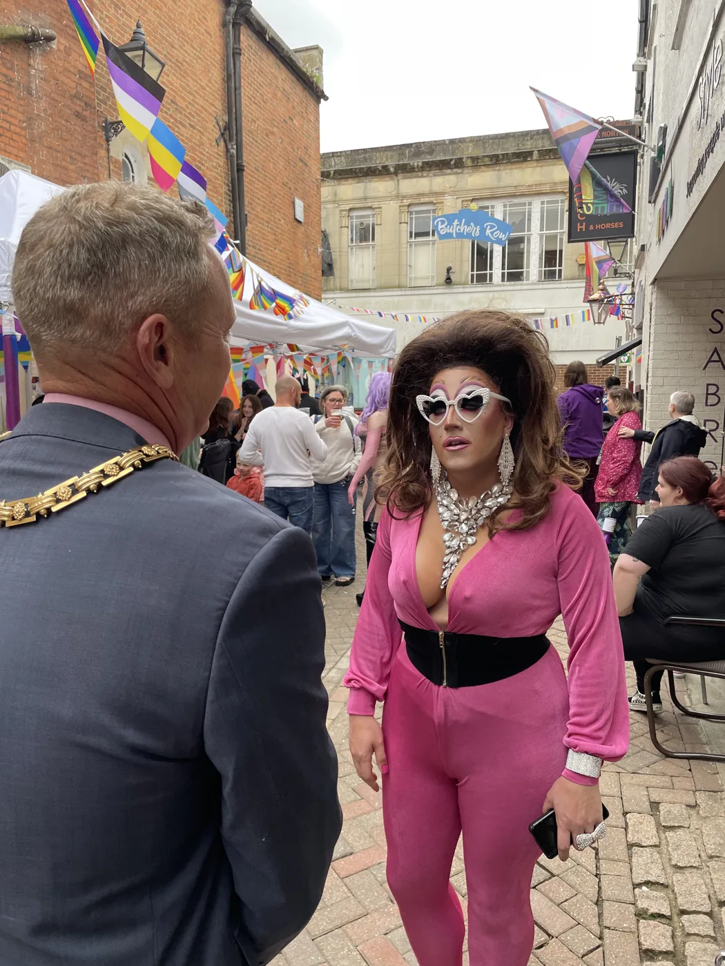 A performer in a pink jumpsuit, big hair and heart-shaped sunglasses talks to a man wearing a mayoral chain on Butchers Row