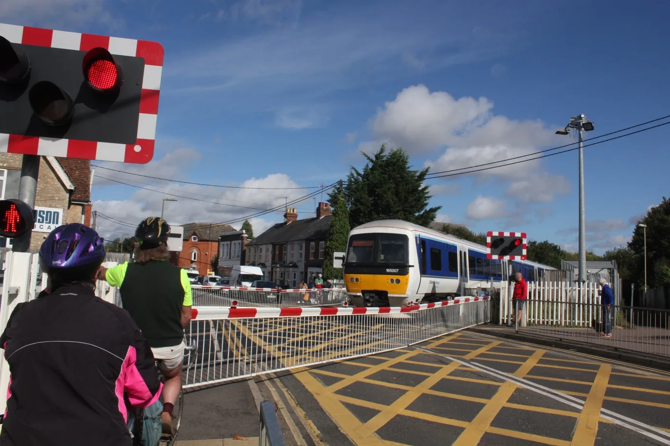 A Chiltern train crosses London Road in Bicester as cyclists wait at the lowered barriers beside Bicester Village station