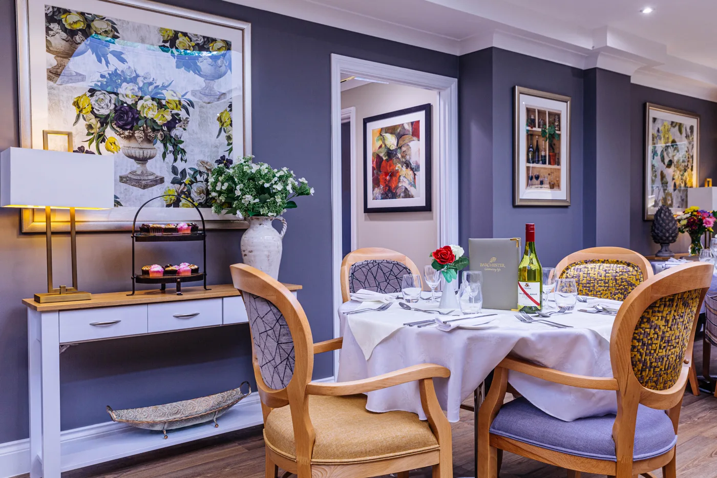 A dining room at Glebefields Care Home with round tables laid with white cloths, upholstered chairs and framed flower paintings on the walls